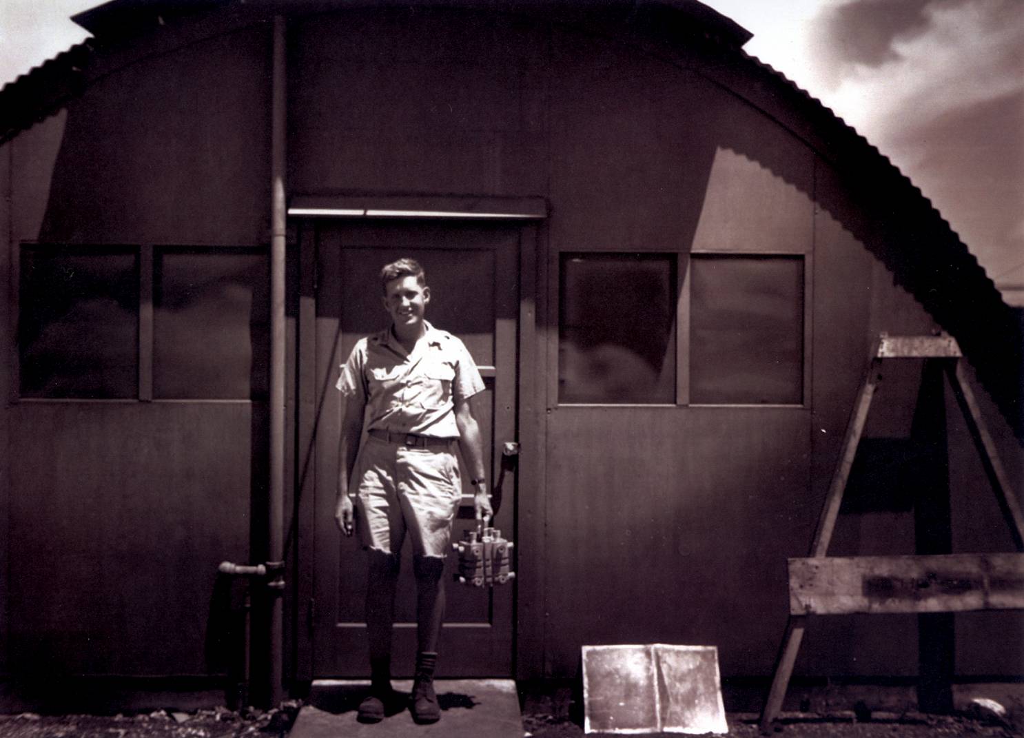 Figure 2 — Harold Agnew on Tinian Island, August 1945, carrying the magnesium plutonium carrying box that held the Nagasaki bomb's pit. The leaning posture suggests roughly 15 pounds of contents — the box its…